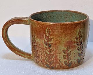 A lovely ceramic mug with a gorgeous textured vine design around the outside and finished with rustic green and blue glazes .